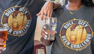 Photo of the torso of two people holding beer glasses wearing dark grey shirts with the 2026 Bull City Beer Challenge design.