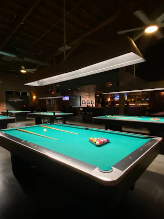 A dark room with six pool tables and a bar in the background.