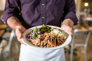 A person in an apron holds a plate of delicious Barbecue from Picnic up close so the camera can get all the details.