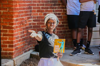 A woman wearing a head scarf and earrings in the shape of Africa performs and holds a brochure that says Baba Chuck Davis.