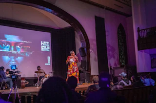 Attendees face the stage as a speaker performs in front of a screen at the Hayti Heritage Film Festival