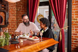 A man and woman in conversation while sitting at a bar. They are smiling and eating.