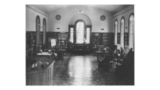 Black and white interior of the library shows wooden floors, bookshelves, large windows and furniture.