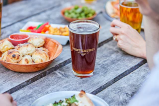 Craft Beverage Summer Editorial Photoshoot with Eric Waters from May 2023.
Close shot of friends drinking beer and eating snacks and pizza on the picnic tables at Ponysaurus.