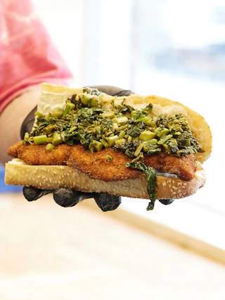 Crispy breaded cutlet sandwich topped with cooked leafy greens on a sesame-seed roll, held in gloved hands.